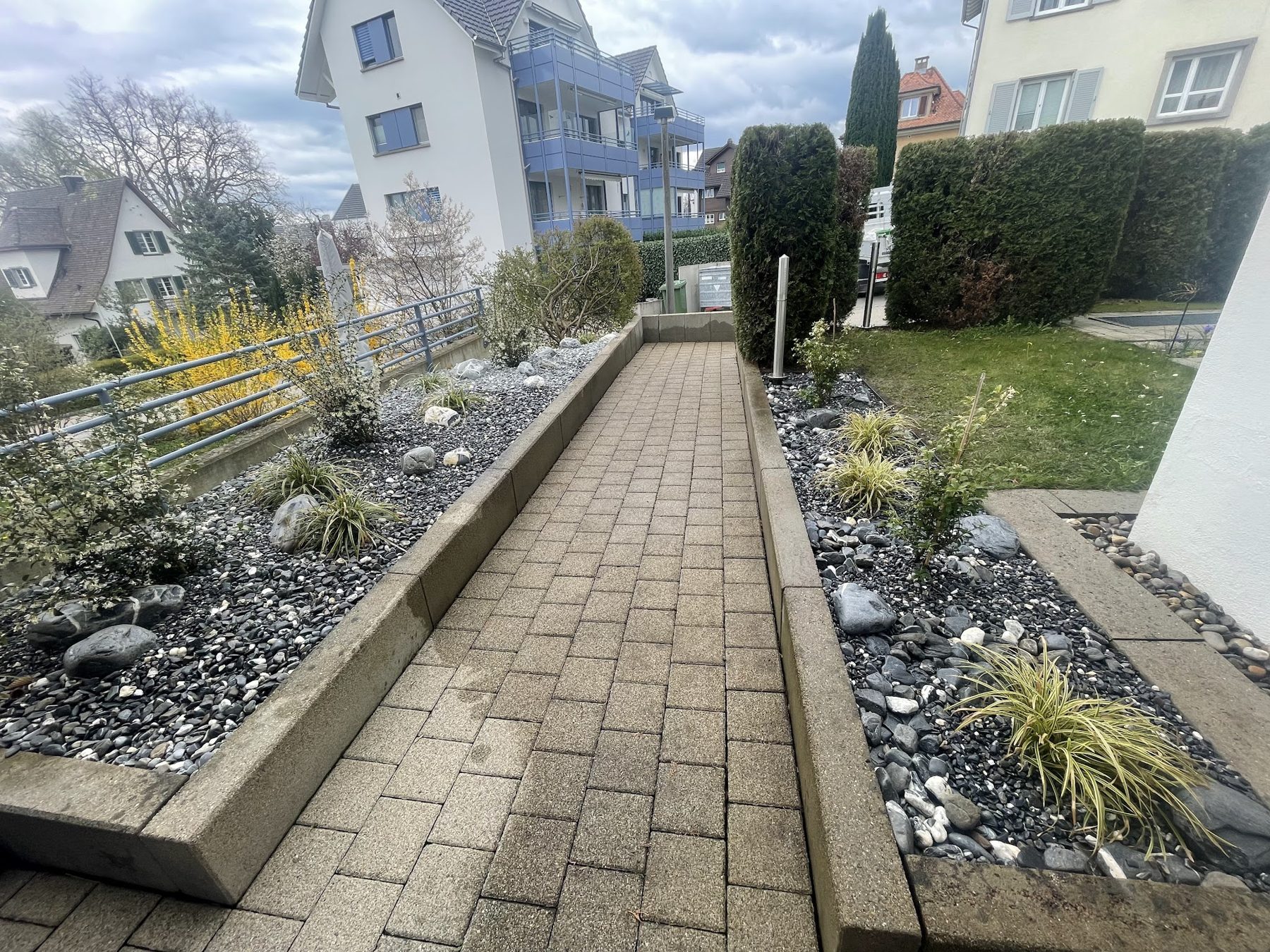 Pflasterweg mit Kiesbeeten, Gräsern und Holzeinfassung bei einem Mehrfamilienhaus